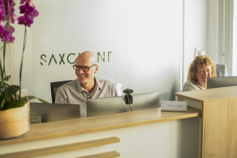 Zwei lächelnde Personen an Schreibtischen in einem modernen Büro, links eine Orchidee, im Hintergrund das Logo 'Saxony' an der Wand."